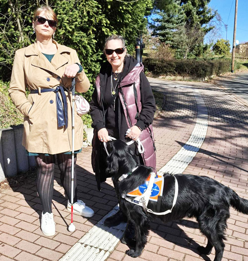 Zwei Frauen stehen auf einem gepflasterten Weg, eine hält einen Blindenstock, die andere hält die Leine eines schwarzen Blindenführhunds mit orange-weißem Geschirr.