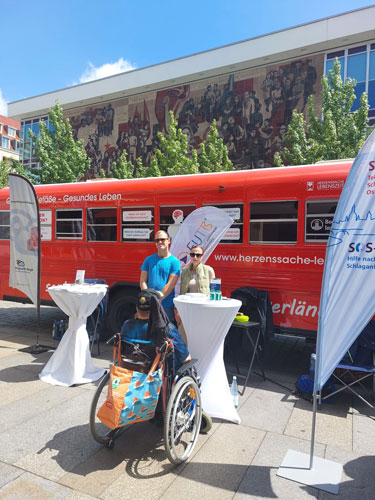 Vor einem großen roten Bus sind Informationsstände mit Beach-Flags und 3 Personen zu sehen, eine davon sitzt in einem Rollstuhl.