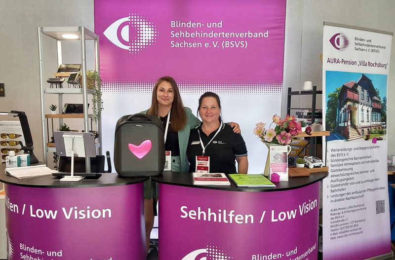 Messestand des Blinden- und Sehbehindertenverbands Sachsen e. V. mit zwei Personen hinter einem lila Tresen mit der Aufschrift 'Sehhilfen / Low Vision' und einem Roll-up mit Informationen zur AURA-Pension 'Villa Rochsburg'.