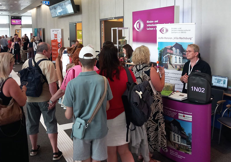 Menschen stehen in einer Schlange vor einem Messestand mit Informationsmaterial und einem Laptop, im Hintergrund weitere Messestände und Besucher in einem hellen Innenraum.