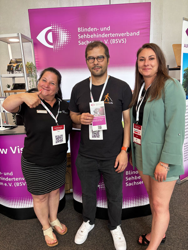 Drei Personen stehen vor einem Messestand mit einem violetten Banner des Blinden- und Sehbehindertenverbands Sachsen e. V. (BSVS), die mittlere Person hält einen Flyer, alle tragen Namensschilder an weißen Bändern um den Hals.