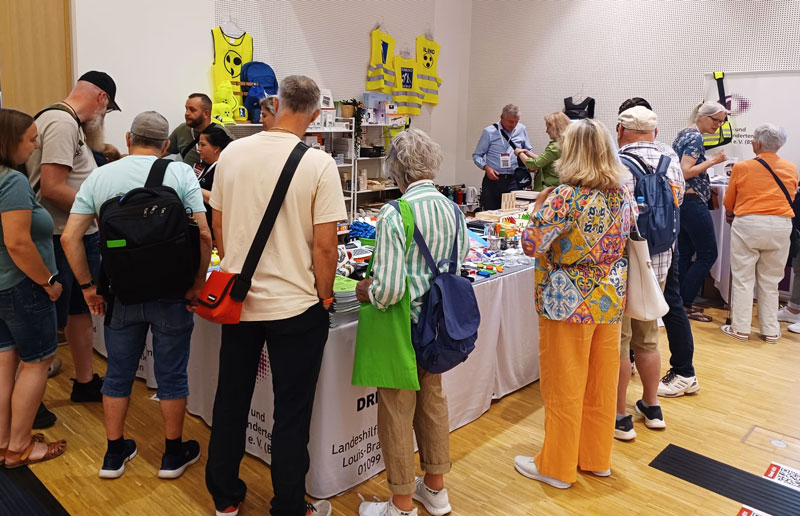 Mehrere Personen stehen an einem Messestand mit Tischen, auf denen verschiedene Produkte und Informationsmaterialien ausliegen, im Hintergrund hängen gelbe Warnwesten an der Wand.