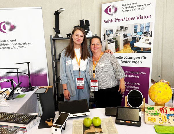 Zwei Frauen stehen an einem Messestand mit Bannern des Blinden- und Sehbehindertenverbandes Sachsen e. V. und dem Thema Sehhilfen/Low Vision, auf dem Tisch liegen elektronische und optische Sehhilfen.