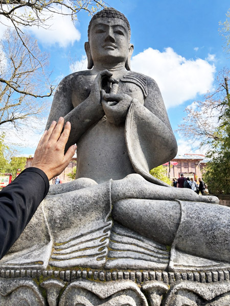 Steinfigur eines sitzenden Buddha mit einer Hand, die versucht, die Figur zu ertasten, vor blauem Himmel und Bäumen.